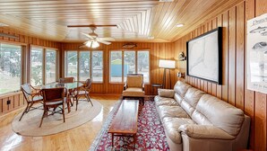 A room with wooden walls, a ceiling fan, a dining table with chairs, a sofa, and large windows. - Exquisite home with private hot tub, game room, & central AC (Salvo)