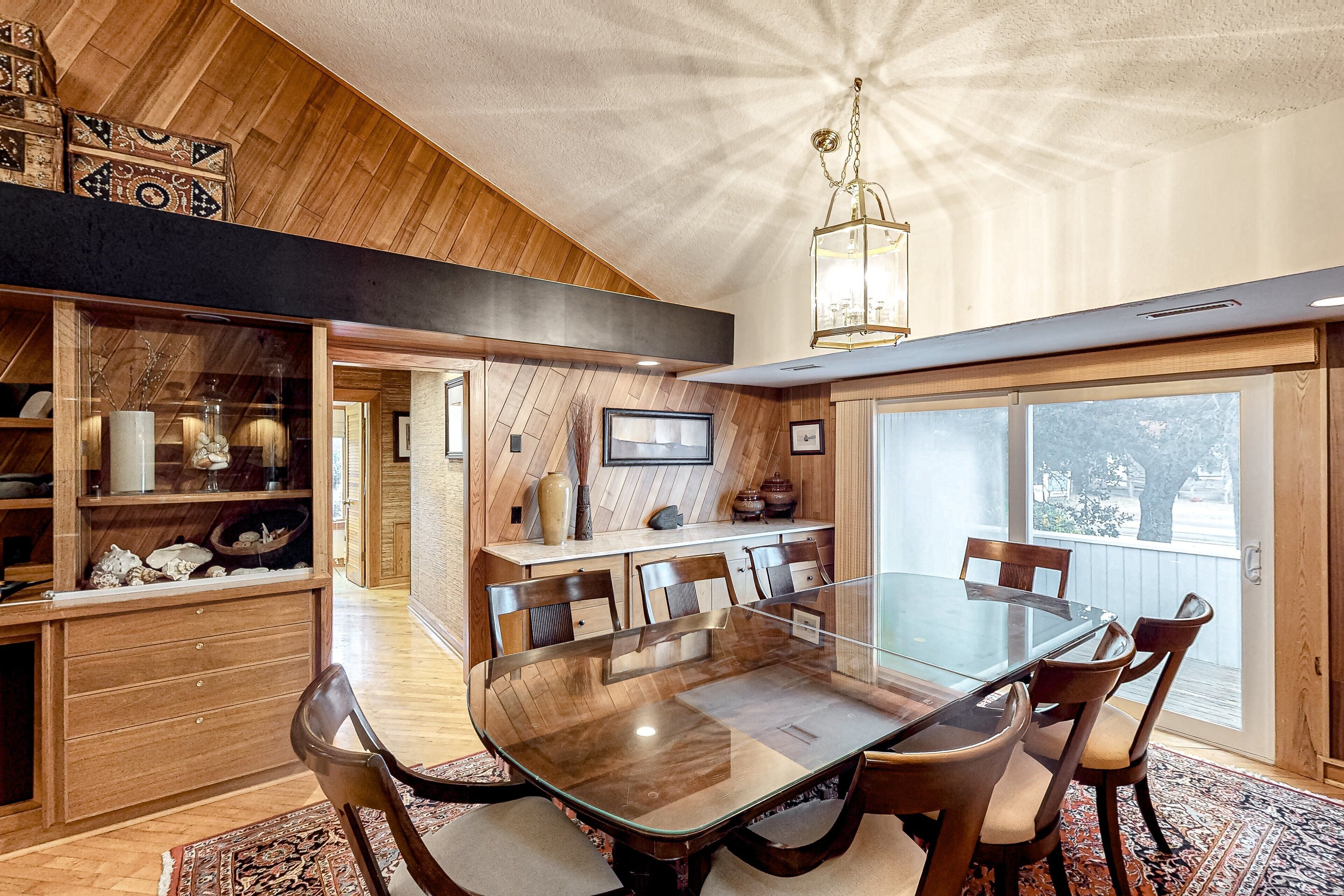 A dining area with a glass-top table surrounded by chairs, a wooden ceiling, and a view through a large window.