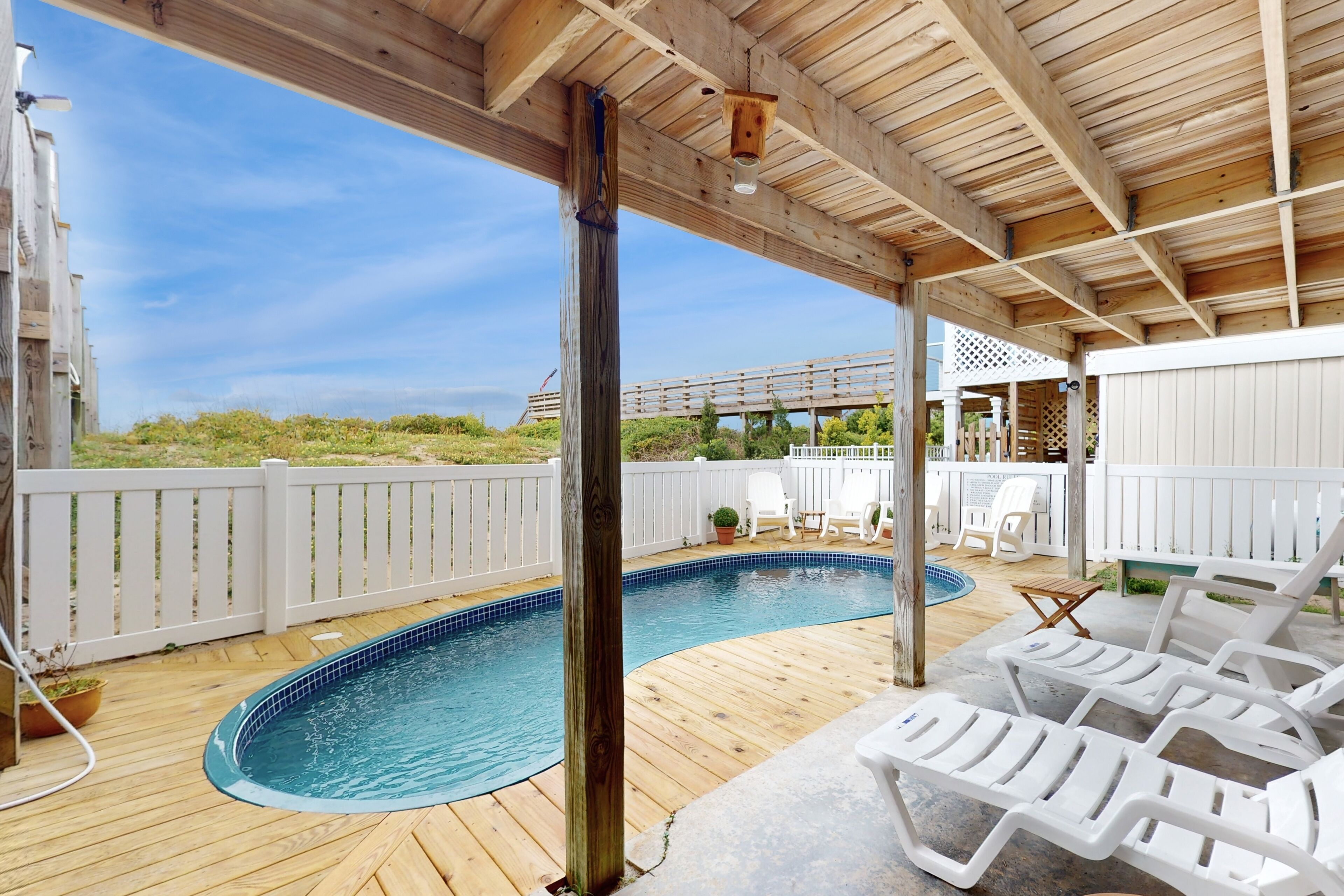 A wooden deck with a swimming pool, white lounge chairs, and a white fence.