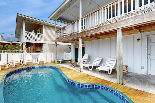A pool area with a wooden deck, white lounge chairs, and a covered deck with a railing.