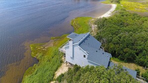 Ett hus vid kusten med en stor terrass med utsikt över vattnet, omgivet av frodig grönska och en sandstrand.