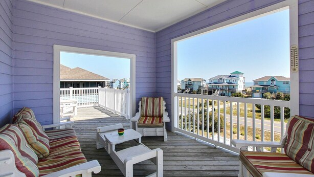 A covered porch with white furniture, including a striped sofa and a chair, and a view of houses in the background. - Outer Banks house with private pool, hot tub, W/D & ocean view - dog-friendly (Avon)