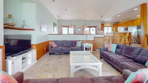 A spacious living room with a flat-screen TV, two sofas, a coffee table, and a kitchen area in the background. - Outer Banks house with private pool, hot tub, W/D & ocean view - dog-friendly (Avon)