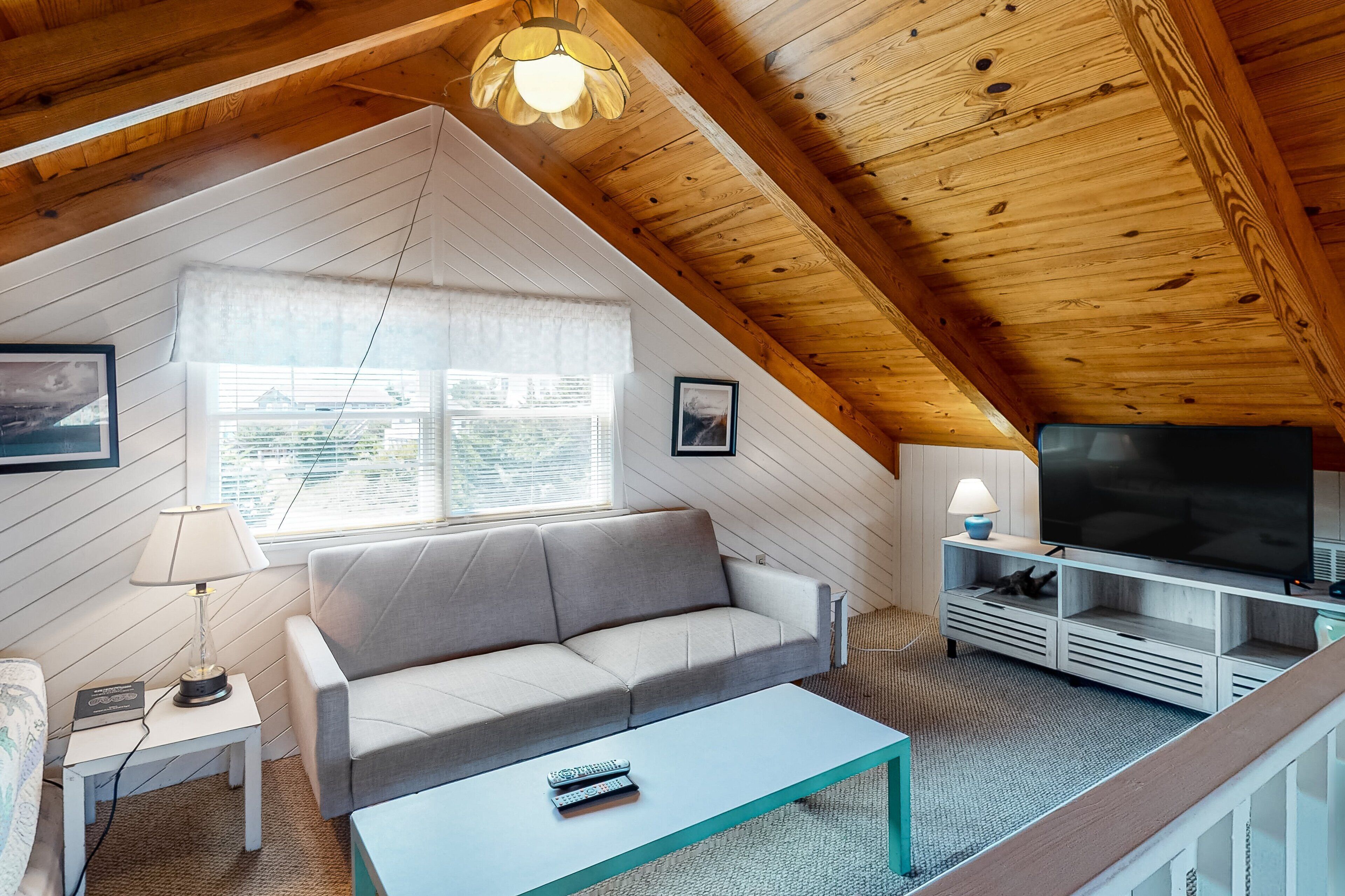 A cozy attic room with a sofa, a coffee table, a TV, and a window with blinds.