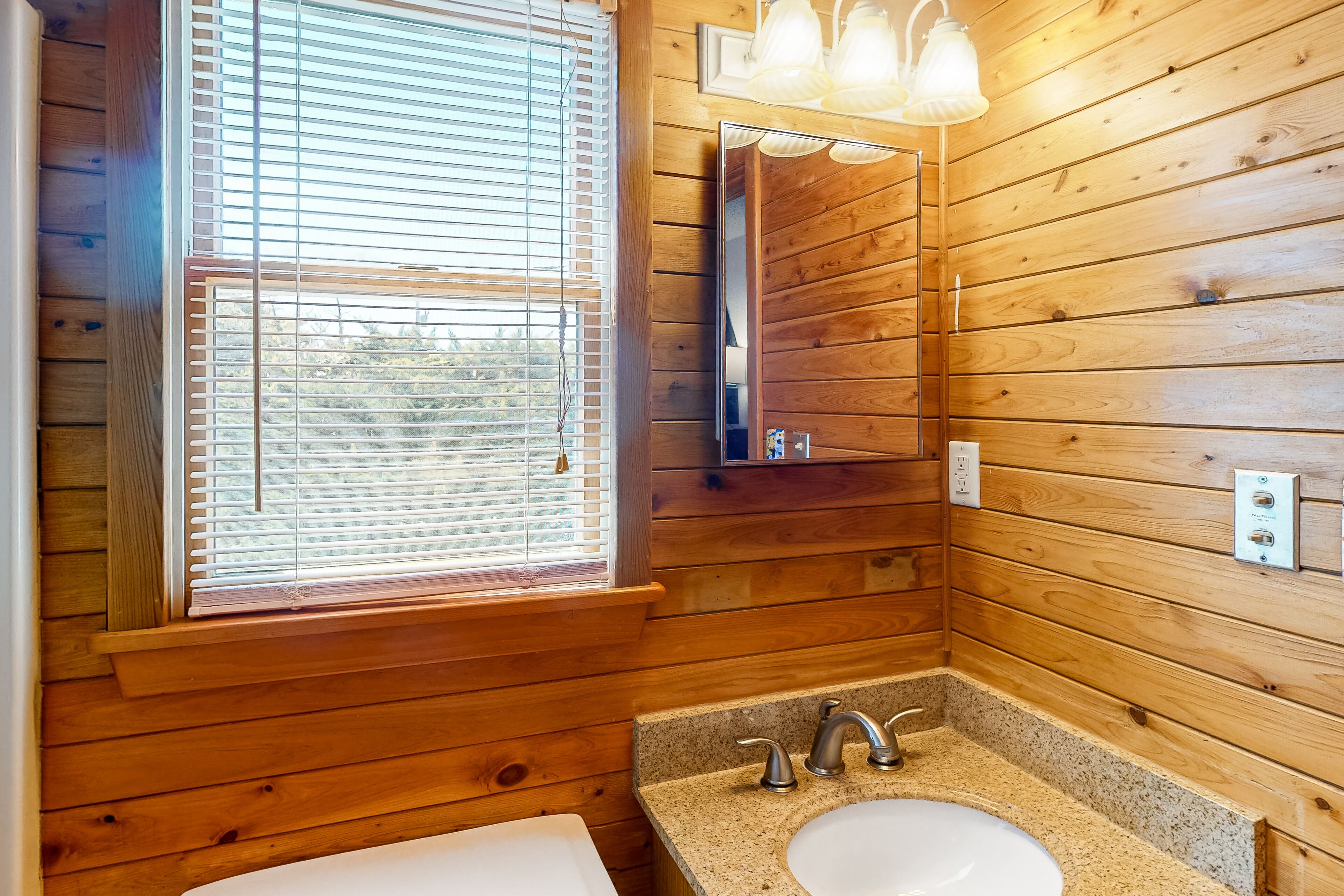 A wooden cabin bathroom with a sink, mirror, and window with blinds.