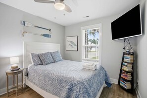A bedroom with a bed, a bedside table with a lamp, a wall-mounted TV, and a bookshelf. - Oak Island Gem: Delightful getaway near the beach with fast WiFi, gas grill, outdoor shower (Oak Island)