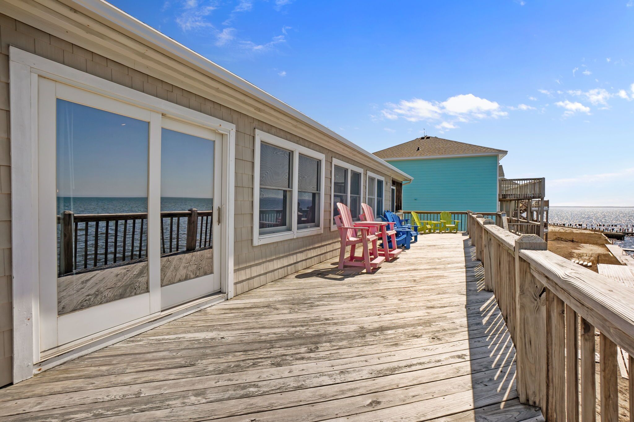 A wooden deck with large windows overlooking the ocean, a blue building in the background, and several colorful Adirondack chairs.