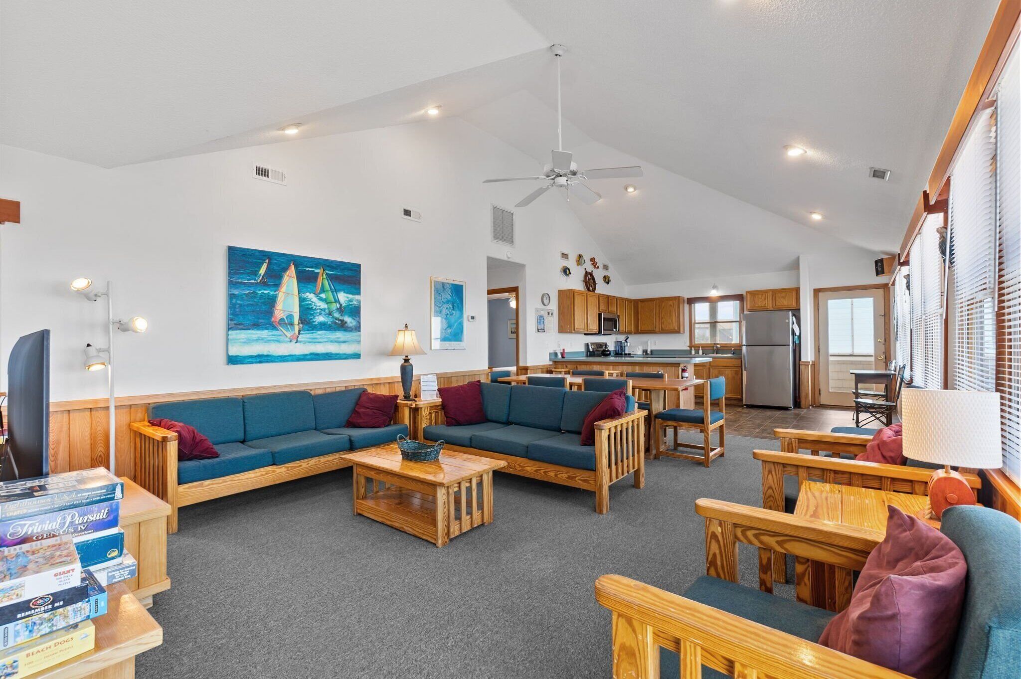 A spacious living area with a sofa, coffee table, and wooden chairs. A kitchen is visible in the background with wooden cabinets and a refrigerator.