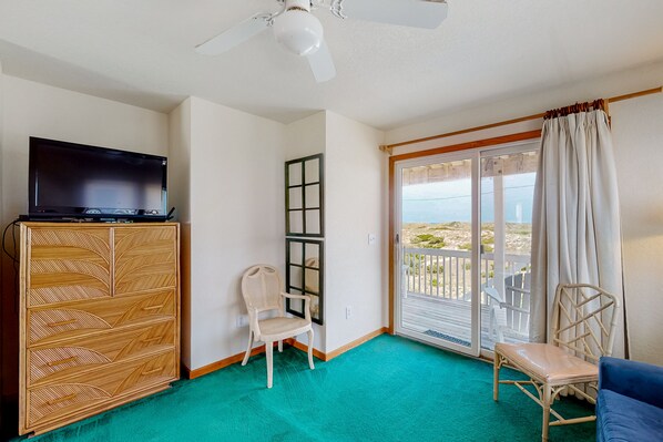 A room with a television on a wooden cabinet, a wicker chair, a blue sofa, and a view of the ocean through a sliding glass door. - Oceanfront home with private pool, hot tub, decks & fast WiFi - dog-friendly (Salvo)