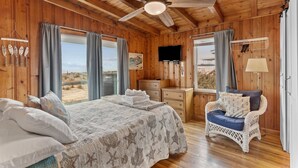 Quarto de cabana de madeira com uma cama, uma cadeira, uma televisão e uma janela com cortinas.