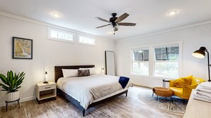 Ein Schlafzimmer mit Bett, Nachttischen, Deckenventilator, Fenster mit Jalousien, einem gelben Sessel und einer Topfpflanze.