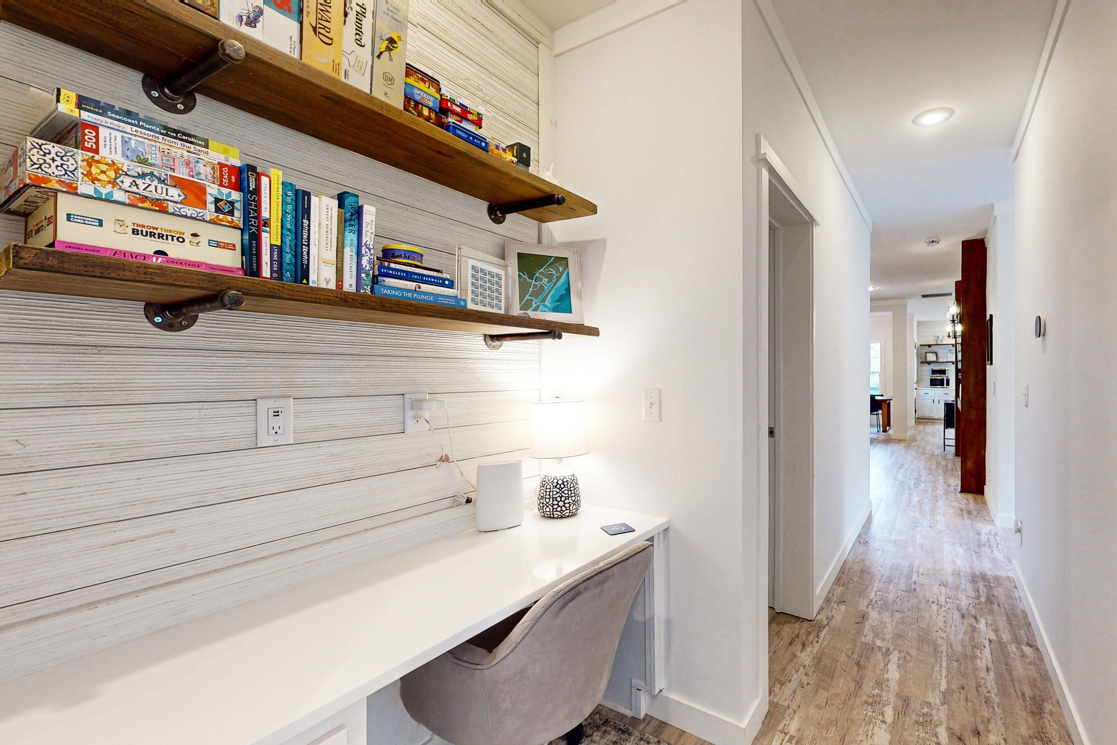 A hallway with a white wall, wooden floor, and a desk with shelves holding books and a lamp.
