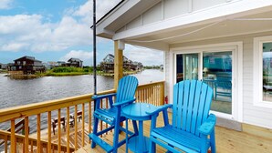 A wooden deck with blue Adirondack chairs and a table overlooking a body of water with houses in the background.
