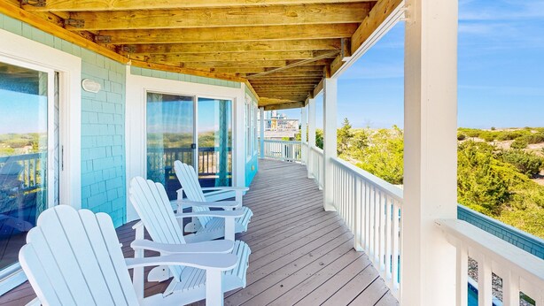 A wooden deck with white Adirondack chairs, a glass door leading to a balcony, and a view of a green landscape. - Multi-level dog-friendly oceanfront home with private pool & hot tub, rec room (Salvo)