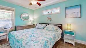 Un dormitorio con cama, ventilador de techo, mesita de noche, una silla y una ventana.