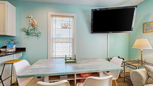 Un área de comedor con una mesa color turquesa, sillas blancas, un televisor montado en la pared y un ventanal con persianas.