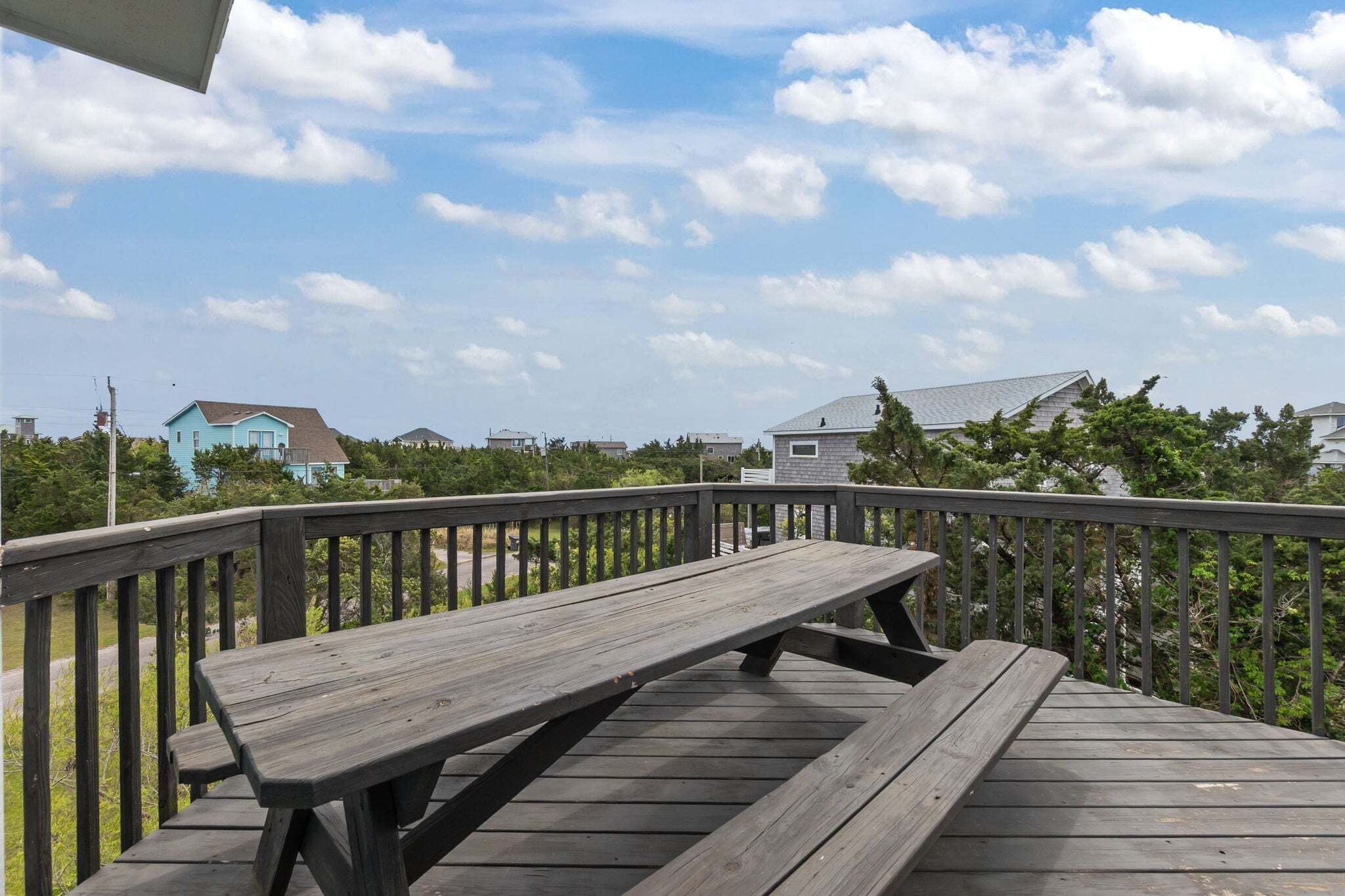A wooden deck with a picnic table, surrounded by a railing, overlooking a residential area with houses and trees.