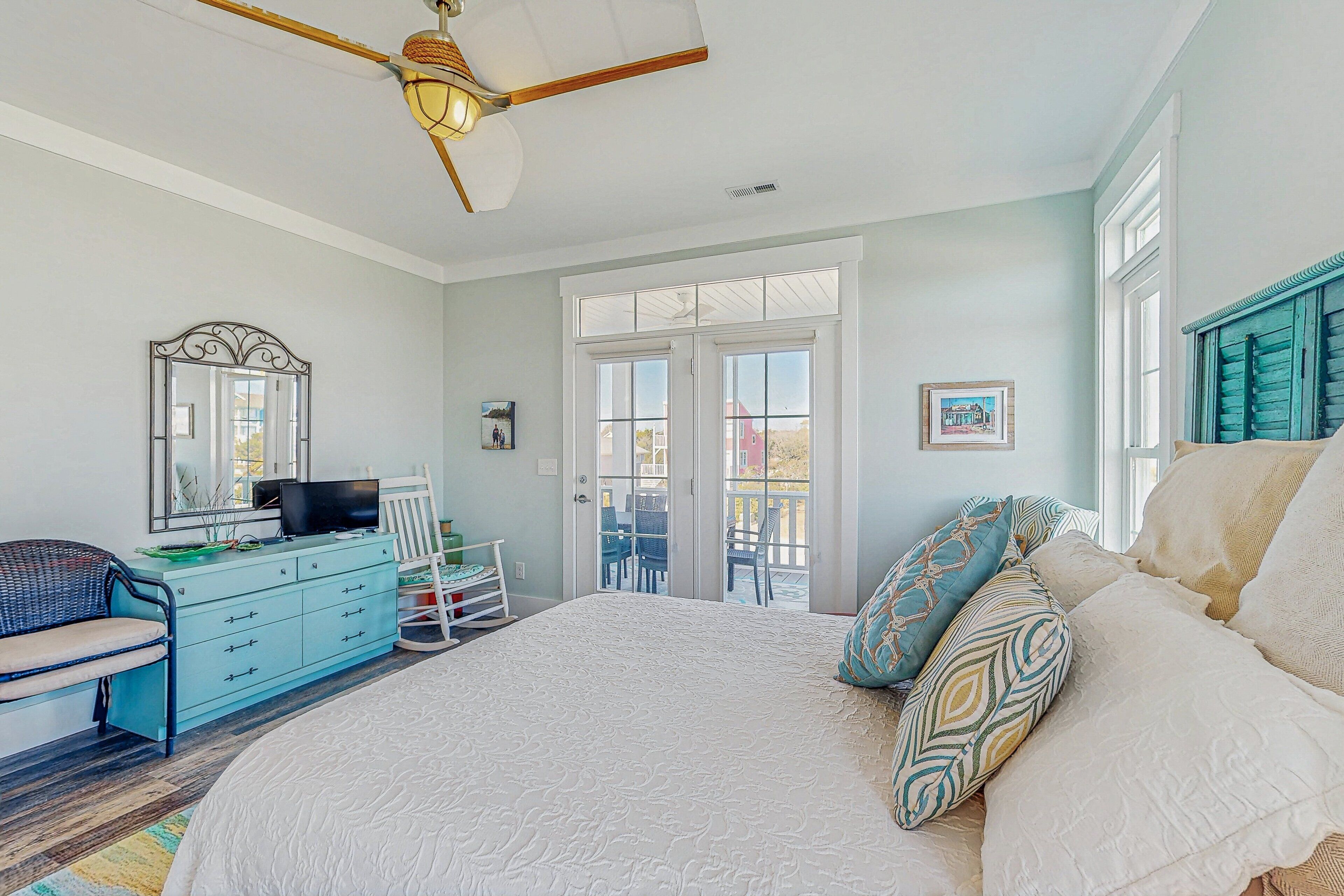 A bedroom with a large bed, a dresser, a television, and a balcony with chairs.