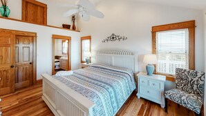 A bedroom with a large bed, wooden floors, a ceiling fan, a window with blinds, and a nightstand with a lamp.