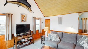 Un salón con techo de madera, un sofá gris, un televisor sobre un mueble de madera y un gran pez montado en la pared.