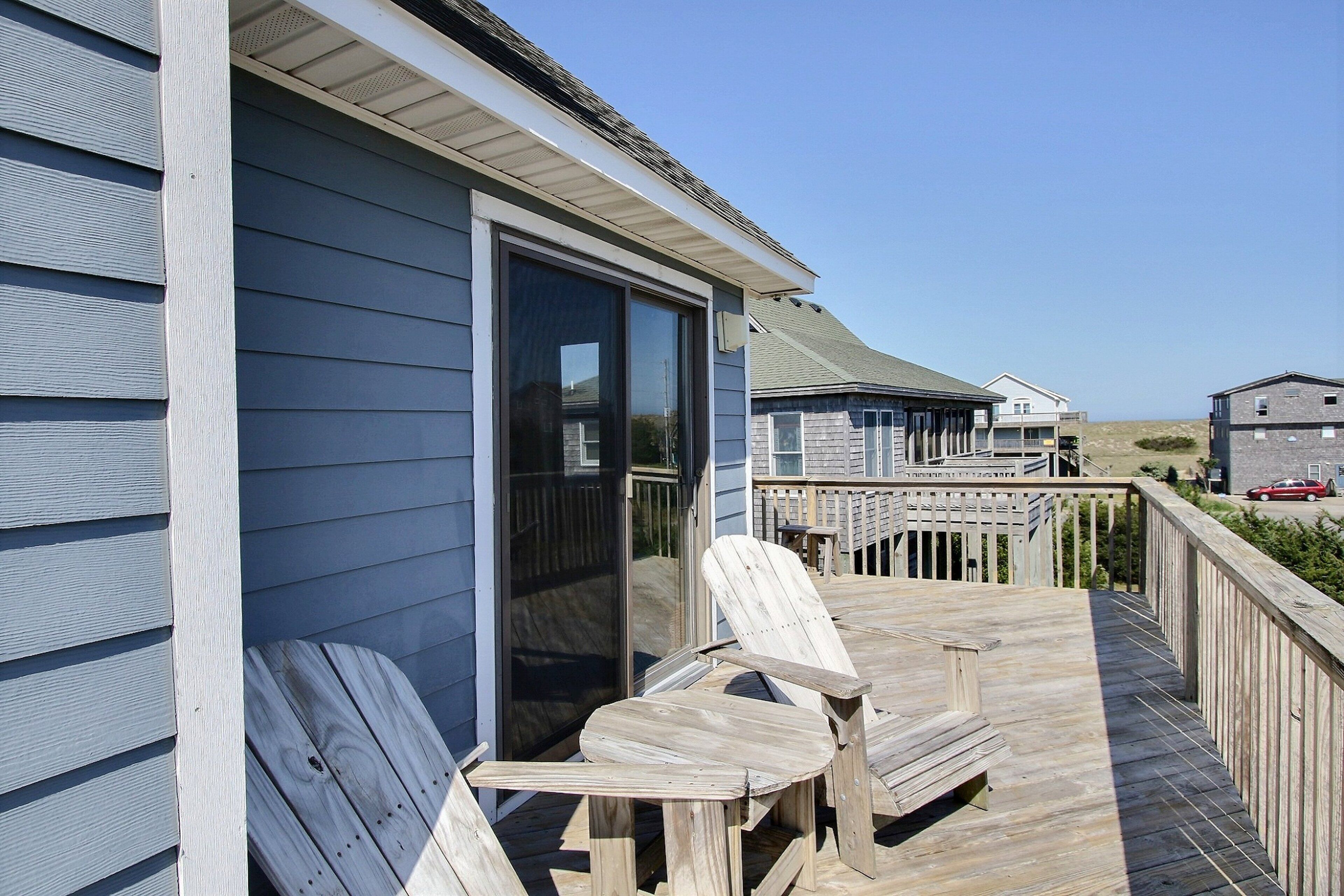 A wooden deck with Adirondack chairs, a glass door leading to a covered area, and a view of the beach and other buildings in the background.