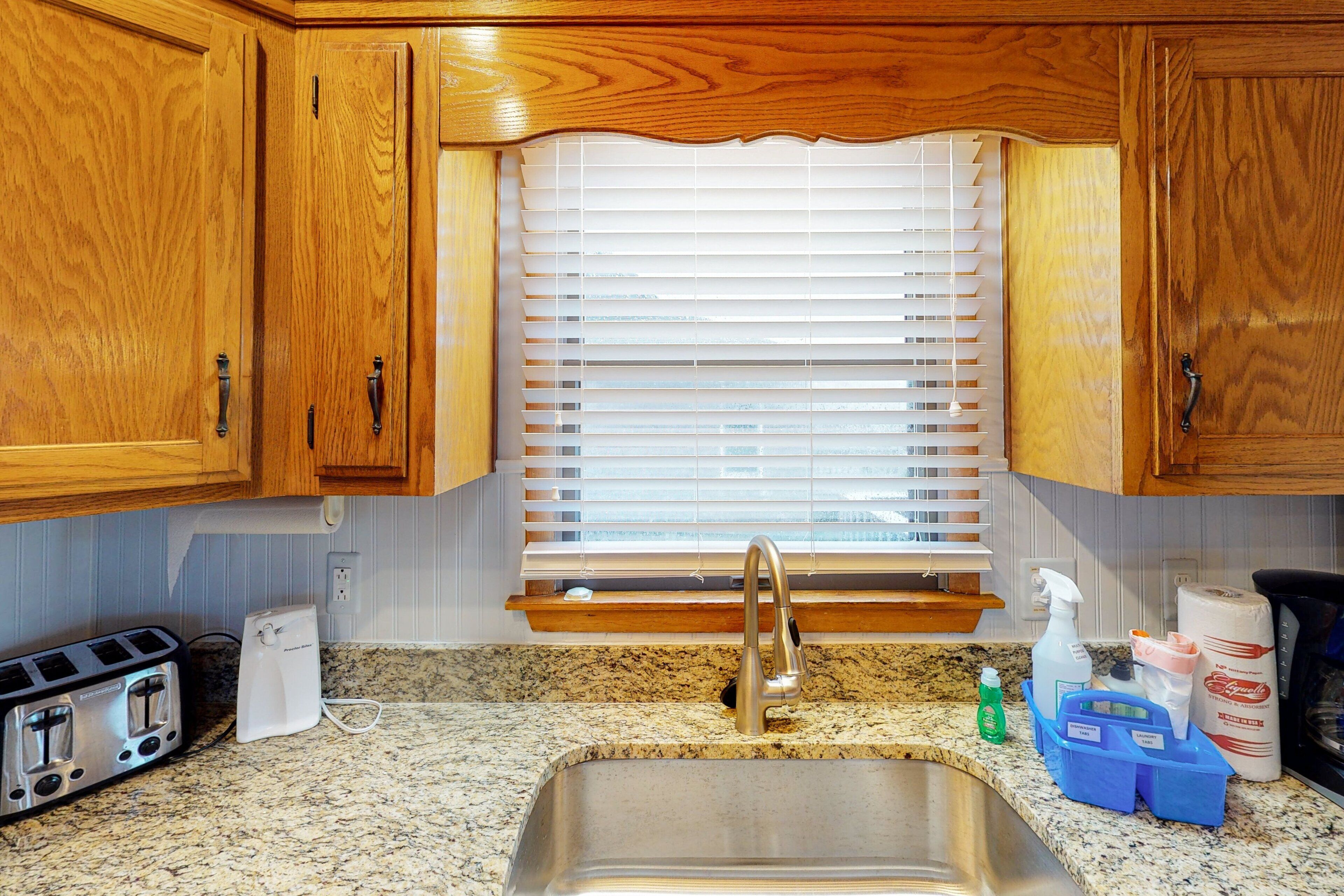 Una cocina con armarios de madera, un lavamanos de acero inoxidable, un tostador, una cafetera y una ventana con persianas.