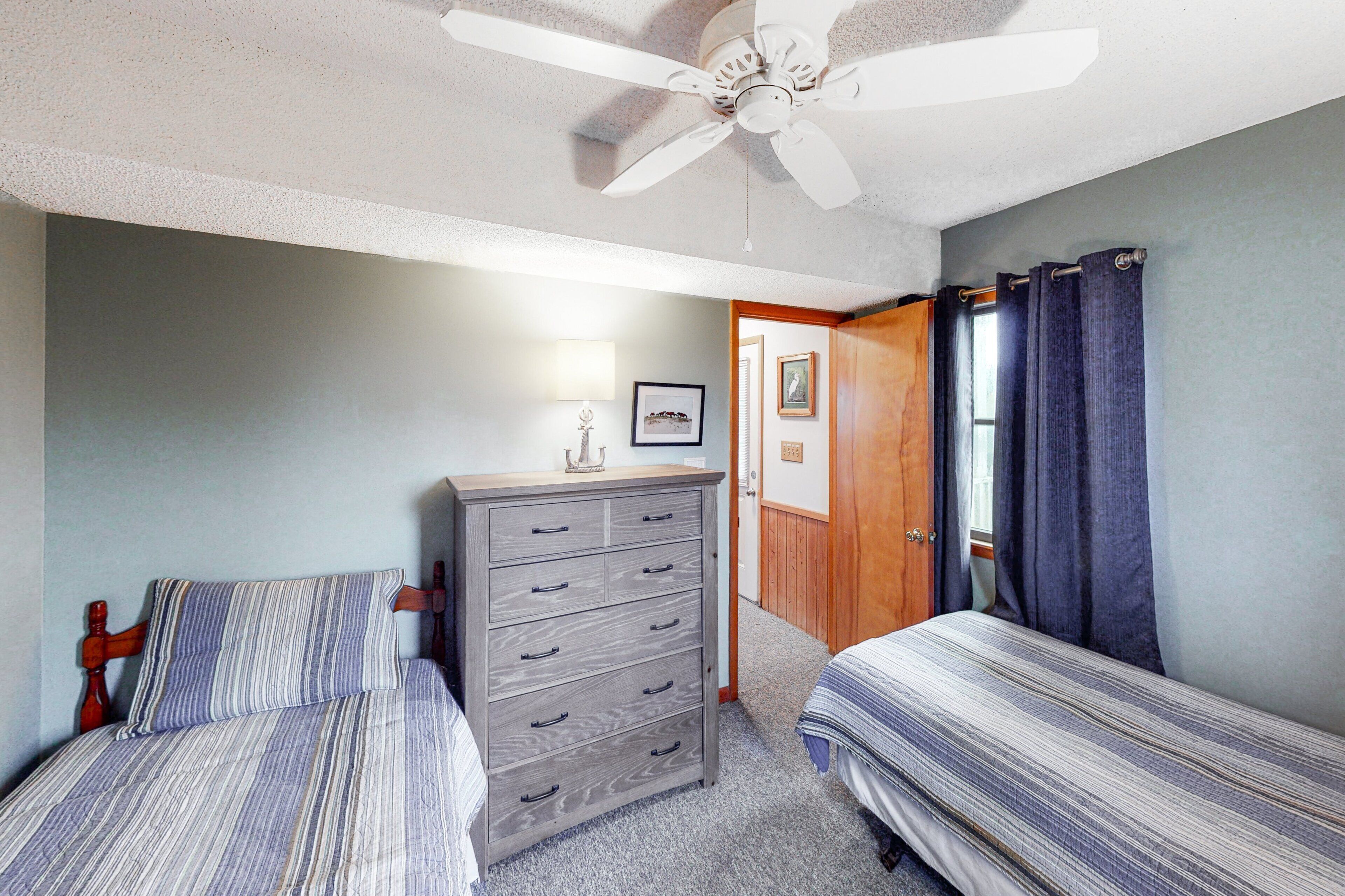 A bedroom with two beds, a dresser, a ceiling fan, and a door leading to another room.