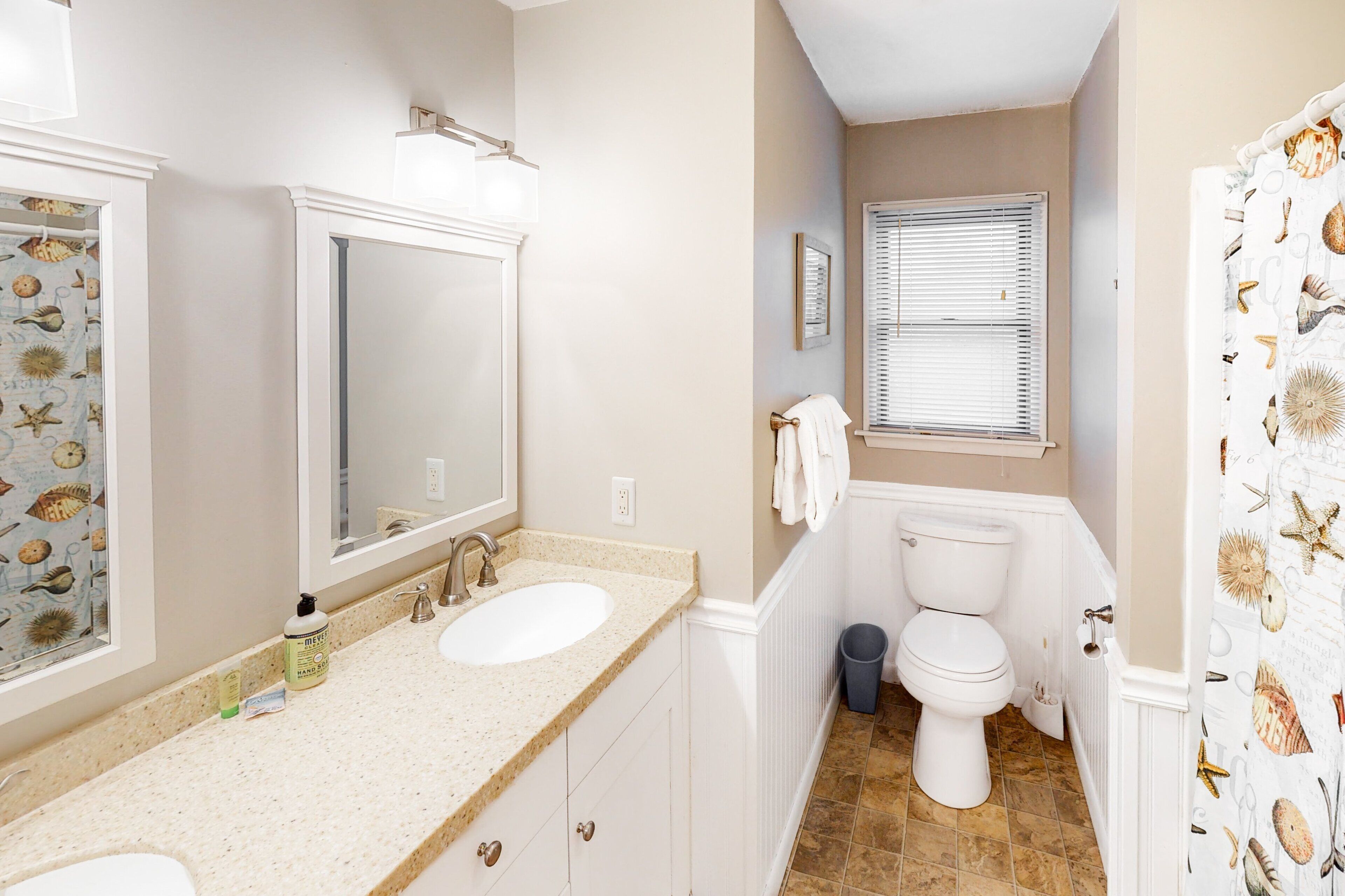 A bathroom with two sinks, a mirror, a toilet, and a window with blinds.
