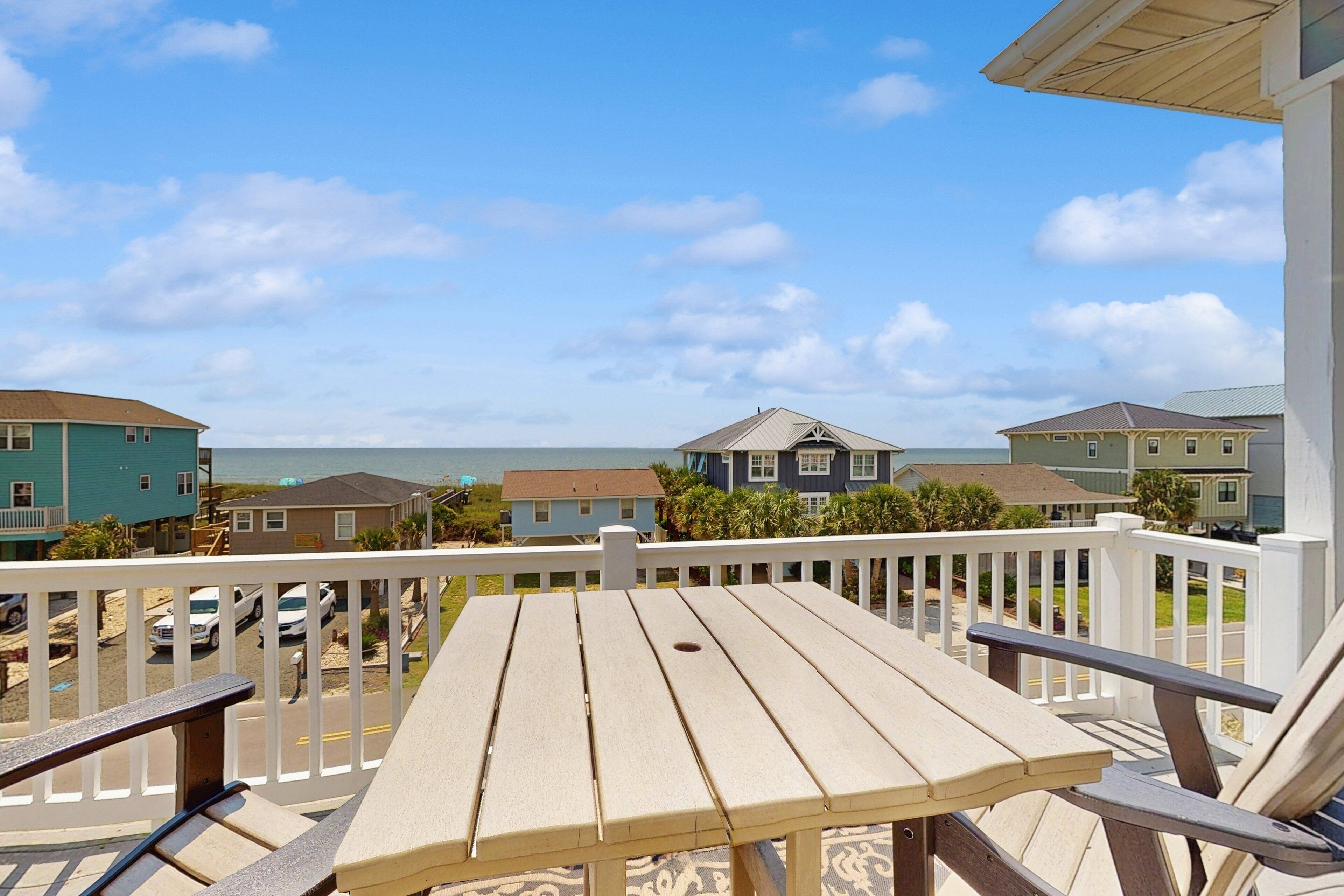Eine Holzterrasse mit Tisch und Stühlen, mit Blick auf einen Strand, im Hintergrund Häuser.