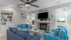 A living room with a fireplace, a ceiling fan, a television, and a bookshelf. - The Doobler: Ocean Views, Near Beach and Golf (Ocean Isle Beach)