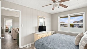 A bedroom with a bed, a dresser, a television, a ceiling fan, and a window with a view of houses.