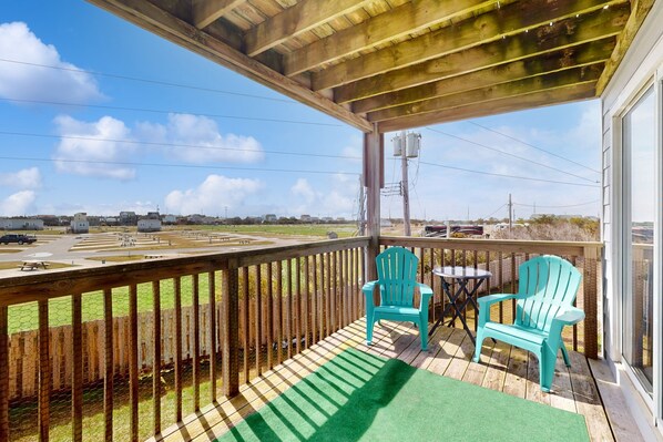 Une terrasse en bois avec deux chaises turquoise et une petite table, d’où l’on aperçoit un parking et des bâtiments éloignés.