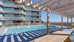 Area piscina con decking in legno e tavoli da picnic, circondata da un edificio a più piani con balconi.