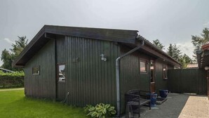 A small, single-story building with a dark green exterior, a sloped roof, and a paved driveway leading to a bench and trash bins. - 4 Star Holiday Home in Vaeggerlose (Vaeggerlose)
