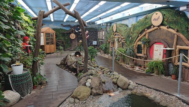 Un sendero de madera que atraviesa un jardín interior temático con un pequeño estanque y cabañas rústicas.