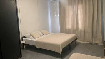 A single bed with a beige blanket, a bedside table, a chair, and a wardrobe in a room with a grey floor and white walls.