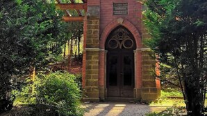Una pequeña capilla de ladrillo con techo puntiagudo, un ventanal circular sobre la puerta y una cruz en la parte superior.
