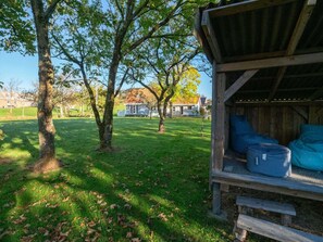 Eine Holzkonstruktion mit Bänken und Taschen, umgeben von einer Rasenfläche mit Bäumen und Häusern im Hintergrund.