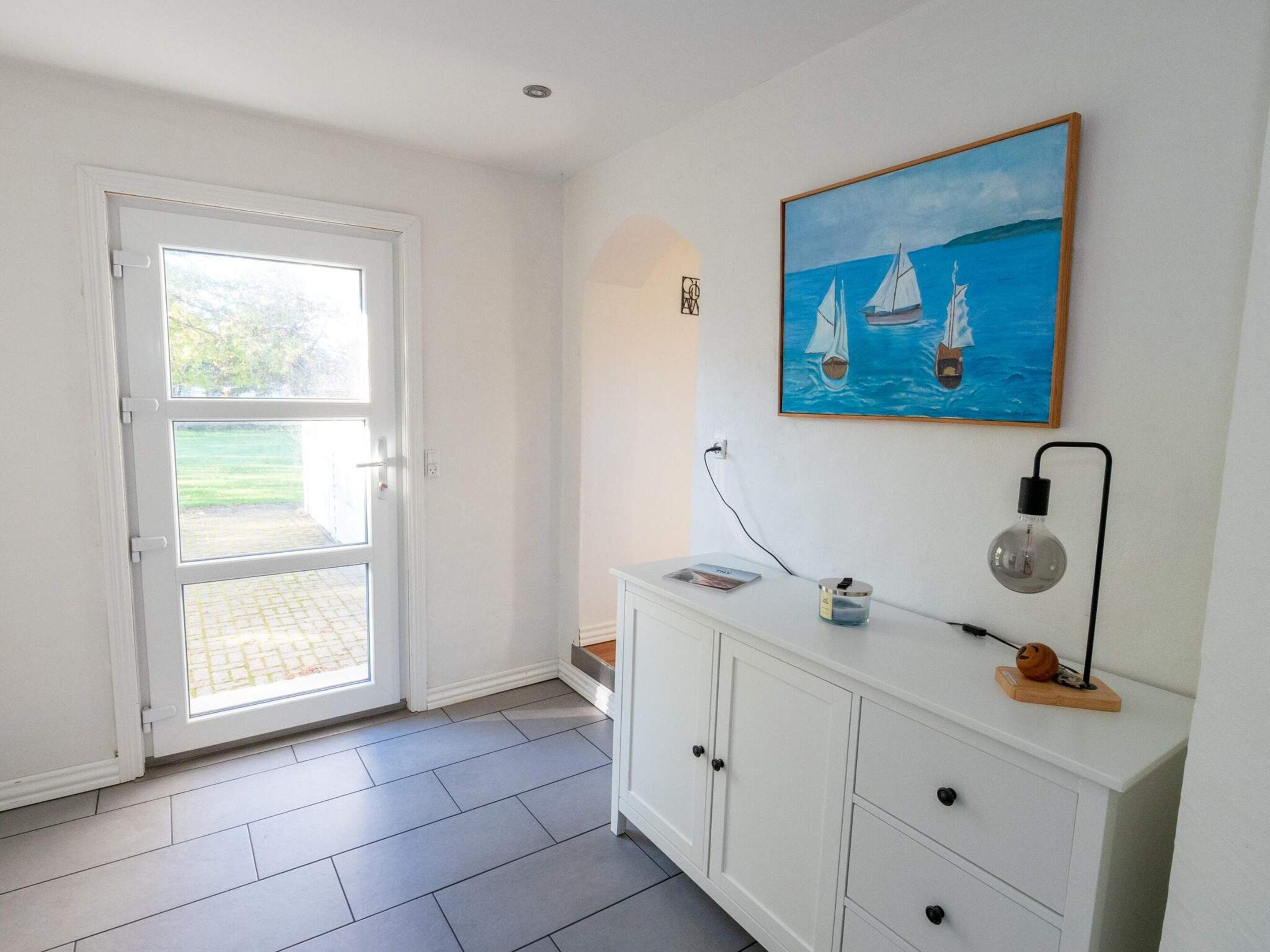 A room with a white door leading to a glass door, a white cabinet with drawers, a painting of sailboats, and a light fixture.