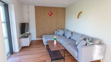 Una sala moderna con un sofá gris, piso de madera, una mesita con un jarrón de flores y un televisor sobre un mueble.