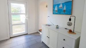 Une pièce avec une porte blanche donnant sur une porte en verre, un meuble blanc avec des tiroirs, un tableau représentant des voiliers accroché au mur et un luminaire.