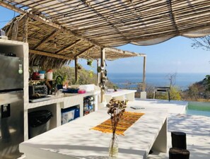 A kitchen with a view of the ocean, a thatched roof, and a white countertop. - Hotel Plaia  (Puerto Angel)