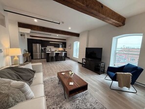 Ein modernes Wohnzimmer mit Sofa, Couchtisch und Fernseher. Im Hintergrund ist eine Küchenzeile mit Kühlschrank und Mikrowelle zu sehen.