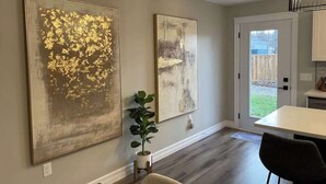 A modern kitchen with a dining area, featuring two large abstract paintings on the wall, a potted plant, and a view of the backyard through the door.