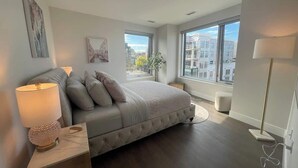 A modern bedroom with a large bed, a bedside table with a lamp, a window with a view of buildings, and a floor lamp. - Downtown, Spotless 2 Bed, 2 Bath with 6 appliances (Halifax)