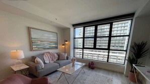 Un salon moderne avec un canapé gris, une table basse en verre, un fauteuil rose et une grande fenêtre donnant sur la ville.