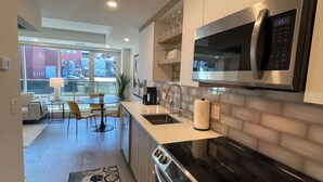 A modern kitchen with a built-in microwave, oven, and sink. There is a dining area with chairs and a table, and a view of the outside through large windows. - Downtown, bright & cozy 1 BR with 6 appliances (Halifax)