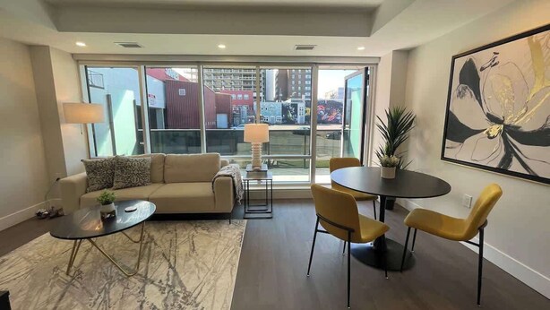 A modern living room with a sofa, coffee table, and chairs, large windows with a city view, and a painting on the wall. - Downtown, bright & cozy 1 BR with 6 appliances (Halifax)