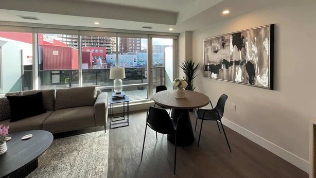 A modern living room with a sofa, a coffee table, a dining table with chairs, a large abstract painting, and a view of buildings through large windows. - Large 1 BR downtown with lots of sunlight (Halifax)