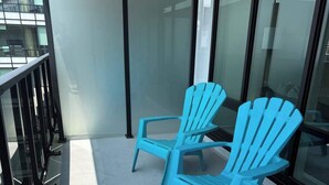 Two blue Adirondack chairs on a balcony with glass railings and a view of a building.
